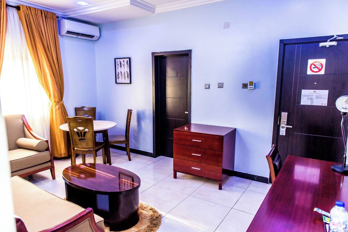 Une chambre d’hôtel comprenant un canapé, une table à manger, une chaise en bois, une petite table ronde, un buffet en bois et une porte sur laquelle est apposé un panneau « interdiction de fumer ».