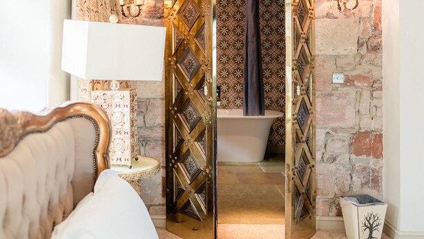 A bedroom with a bed, a nightstand, a lamp, a bathroom with a bathtub, and a stone wall. - Newton Hall Courtyard Rooms (Alnwick)