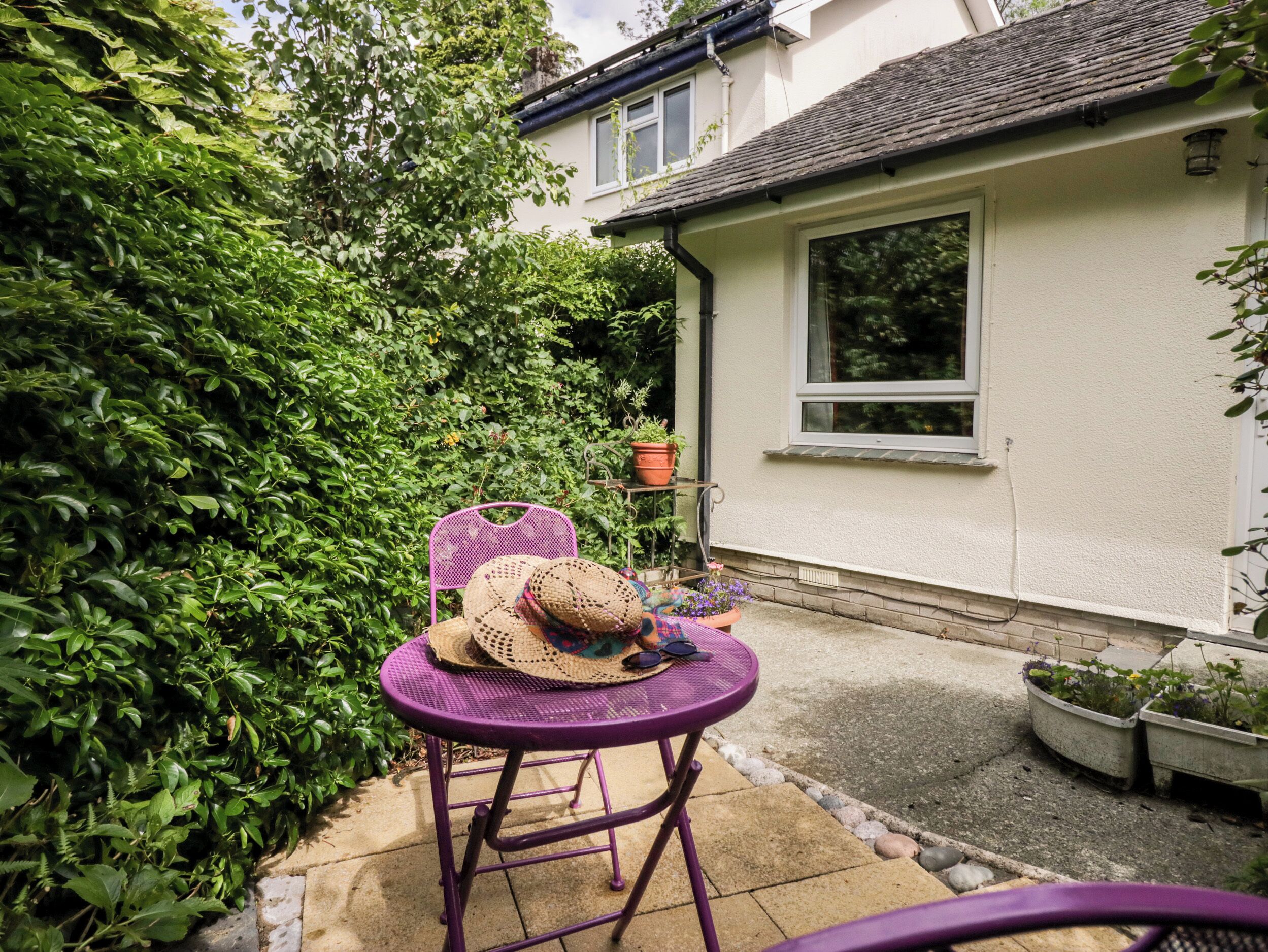 A patio with a purple metal table and chairs, surrounded by lush greenery and a white house with a window.