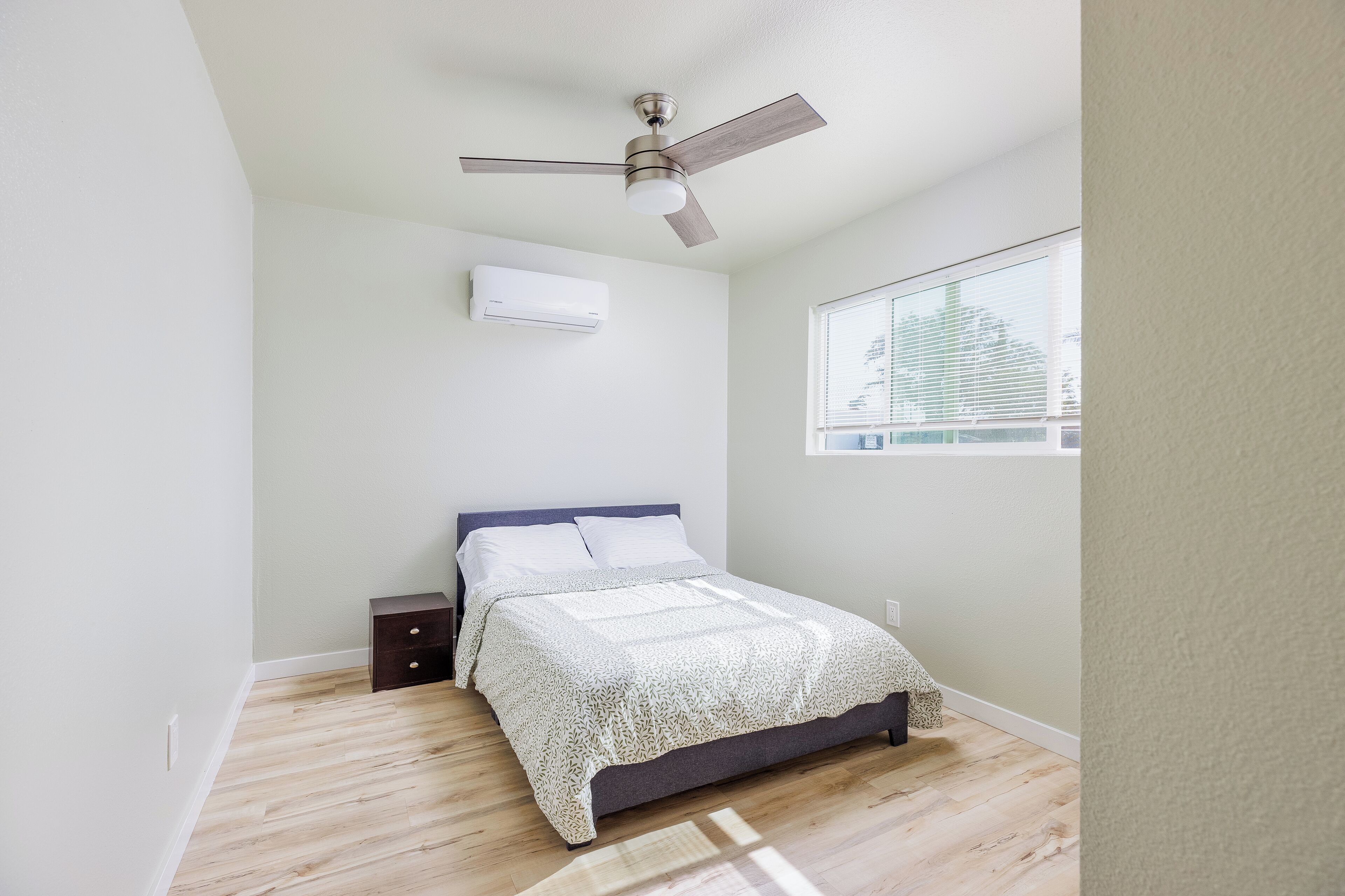 A bedroom with a bed, a ceiling fan, an air conditioner, a window, and a nightstand.