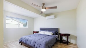 Een slaapkamer met een bed, een plafondventilator, een airconditioning en een raam, plus een nachtkastje.
