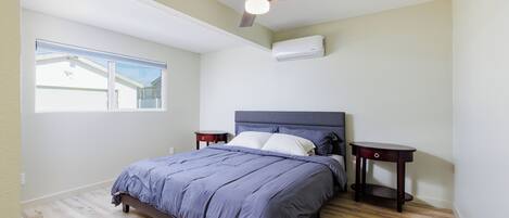 Een slaapkamer met een bed, een plafondventilator, een airconditioning en een raam, plus een nachtkastje.