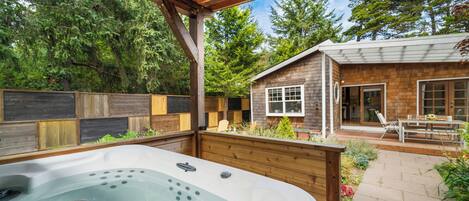 A hot tub under a wooden pergola in a backyard with a wooden fence, a wooden deck, and a house with a window and door.