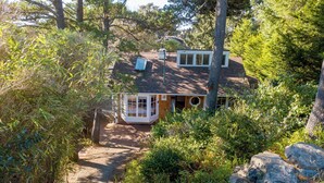 Une maison entourée d’une végétation luxuriante et d’arbres, avec un porche bien visible et un chemin menant à l’entrée.
