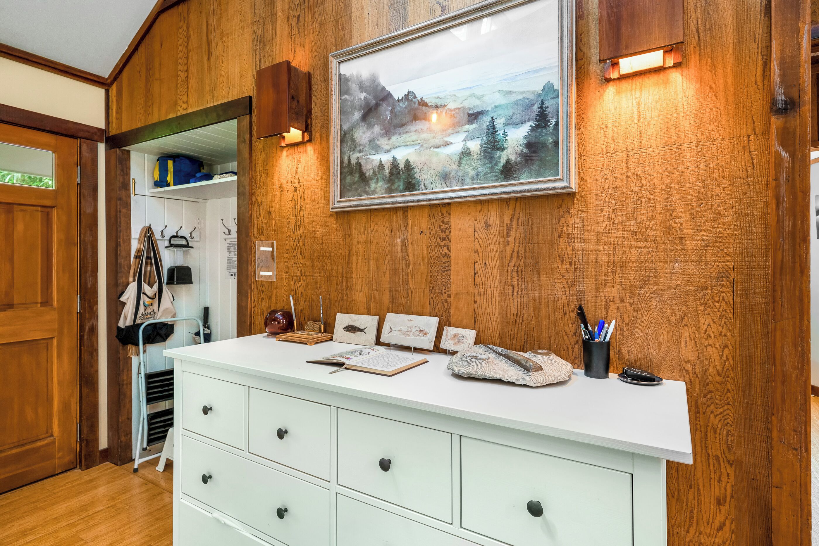Ein Zimmer mit Holzwänden, einer weißen Kommode, einem gerahmten Landschaftsbild und einem kleinen Schreibtisch mit einer Lampe und einem Stiftekännchen.