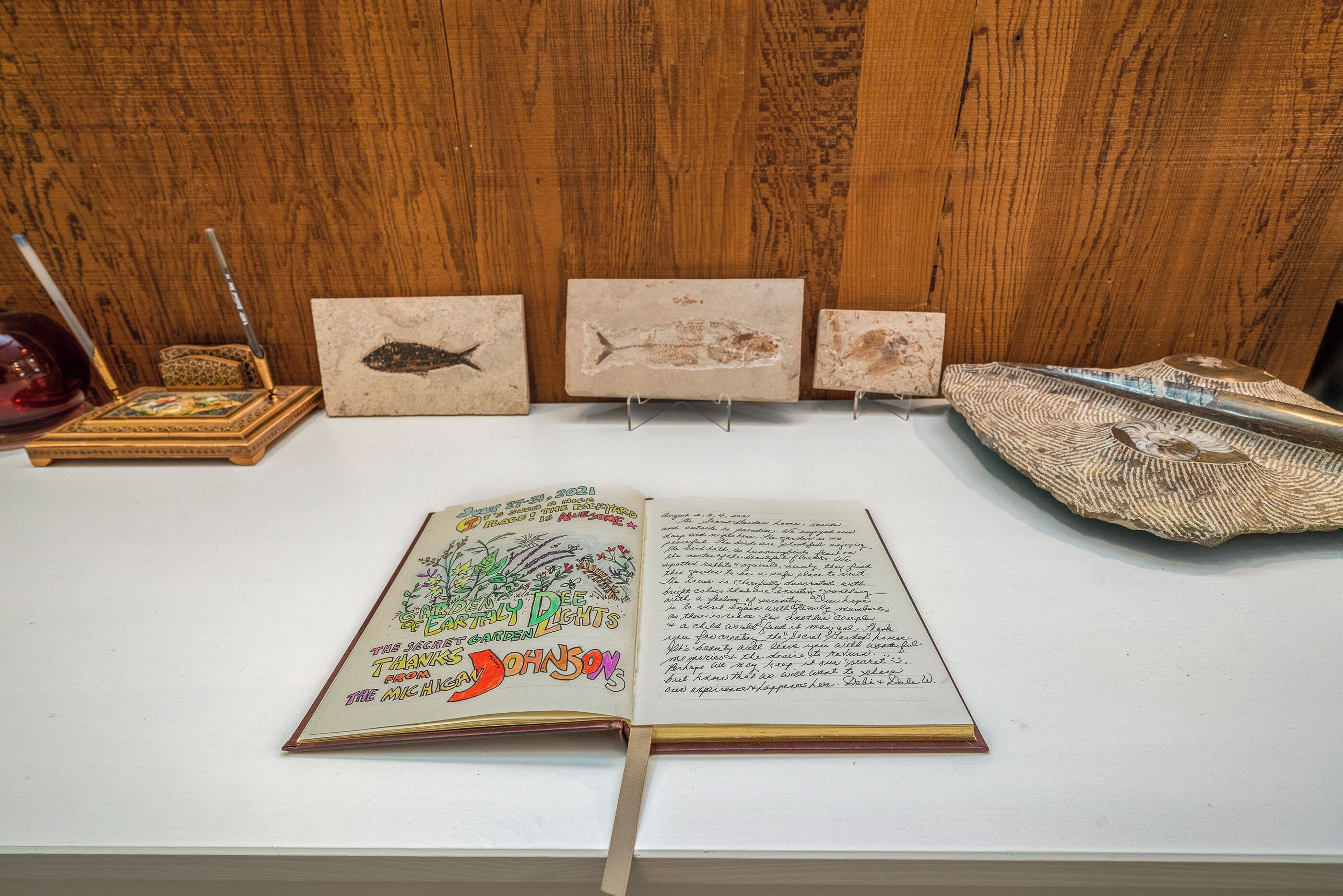 An open book with handwritten notes on a white surface, surrounded by three framed fish paintings and a pair of scissors.