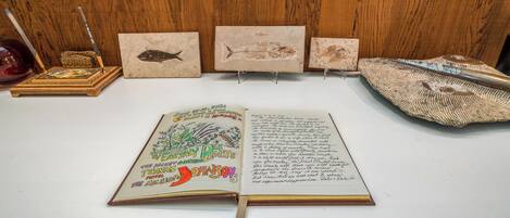 An open book with handwritten notes on a white surface, surrounded by three framed fish paintings and a pair of scissors.