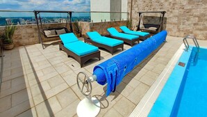 Un espace piscine sur le toit, avec des chaises longues et une chaise suspendue, une piscine recouverte d  ’une bâche bleue, et une vue sur la ville en arrière-plan.