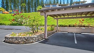 A parking area with a covered walkway, stone planter with flowers, and a backdrop of trees and hills.