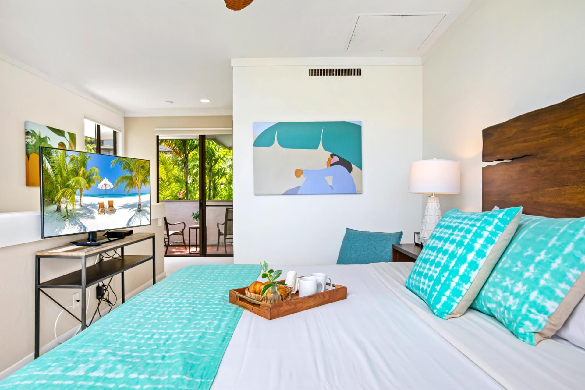 Una camera d'albergo con un letto, una scrivania con un computer, una TV che mostra una spiaggia e un balcone con vista su palme.