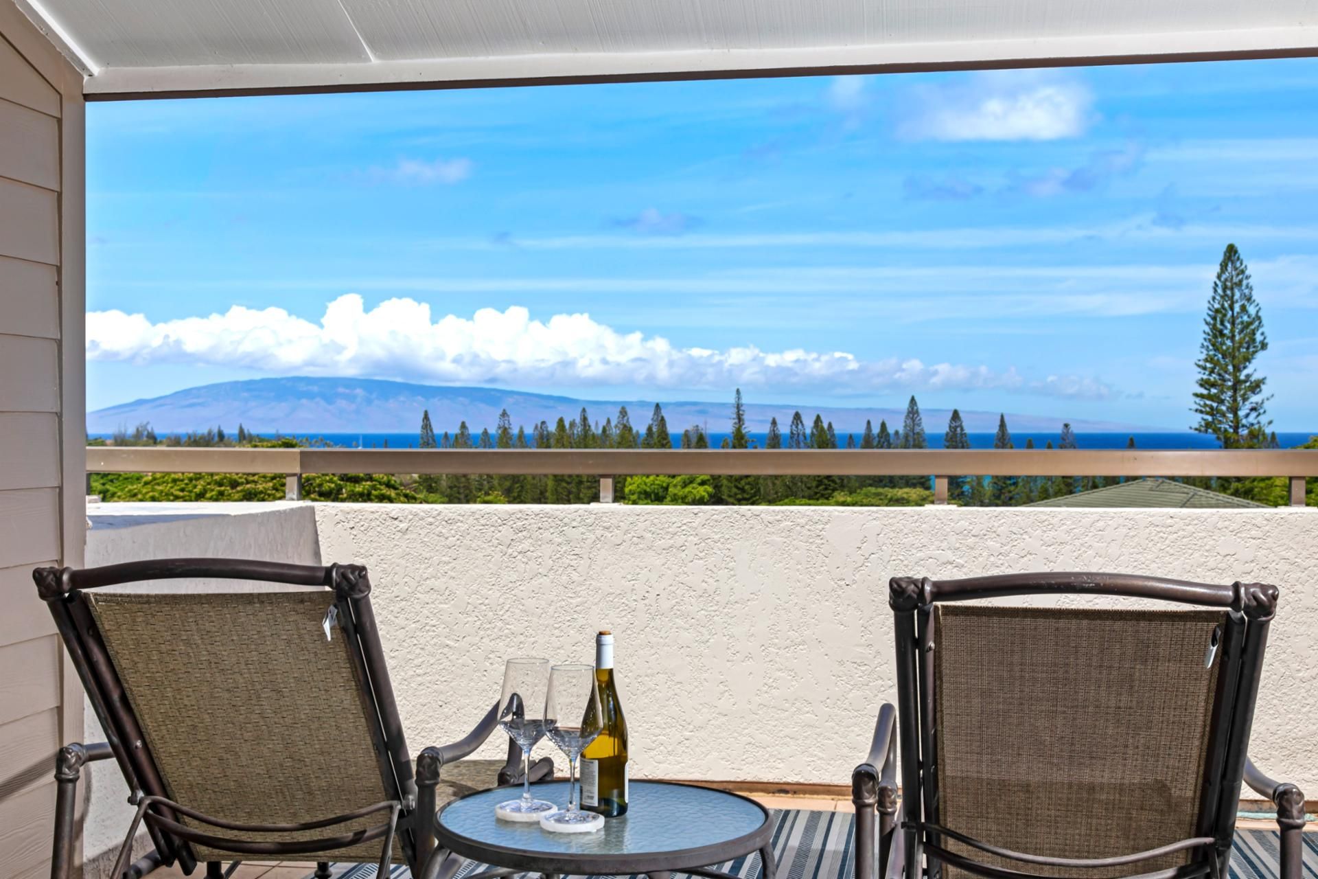 A balcony with two wicker chairs, a small round table, and a bottle of wine. The view includes a mountain and the ocean.