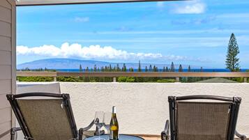 A balcony with two wicker chairs, a small round table, and a bottle of wine. The view includes a mountain and the ocean.