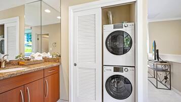 A laundry room with two stacked front-loading washing machines and a dryer, a countertop with a sink and toiletries, and a mirror reflecting the room.