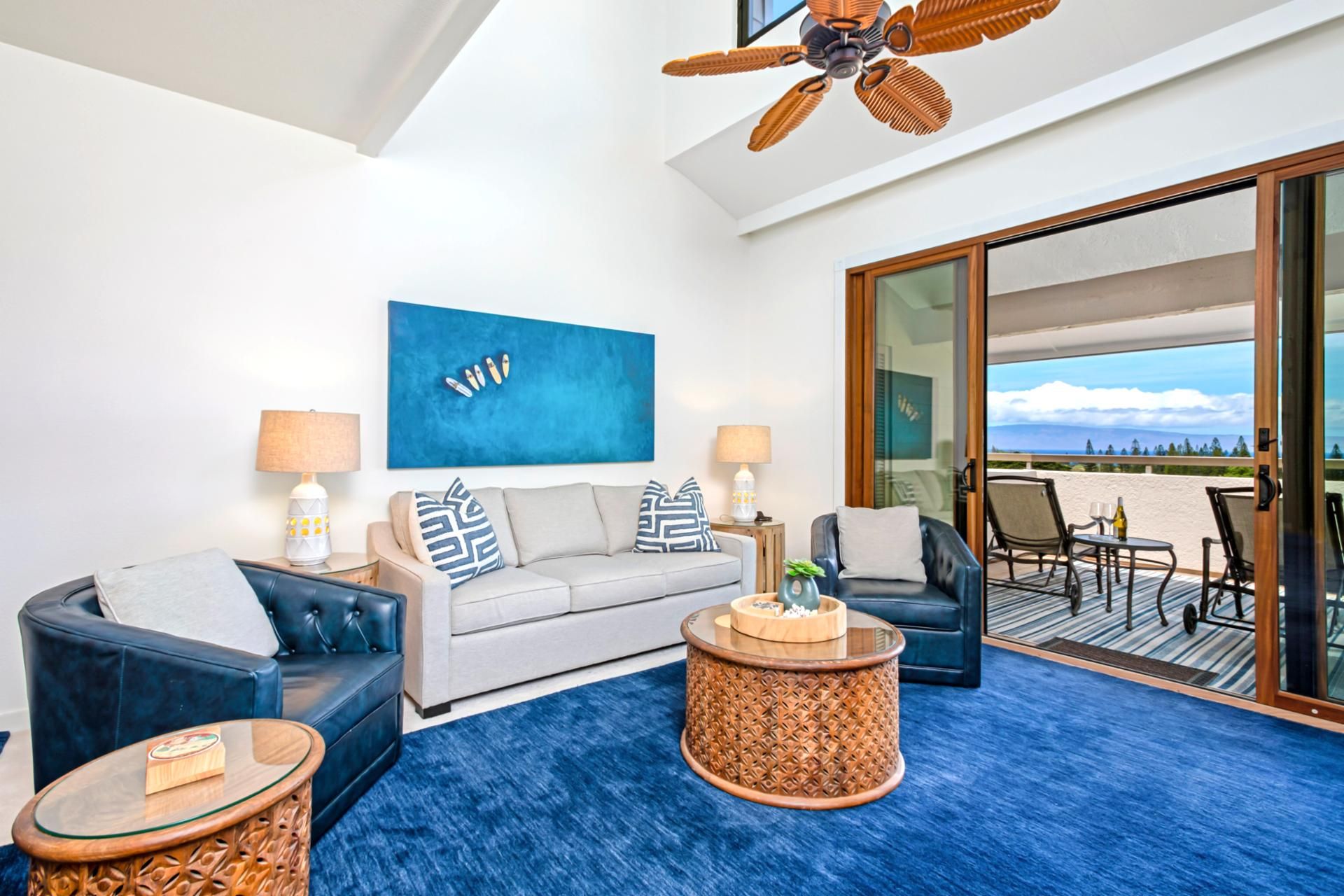 Un salon moderne comprenant un canapé, deux fauteuils, une table basse et un ventilateur de plafond. Une grande fenêtre offre une vue sur la plage.