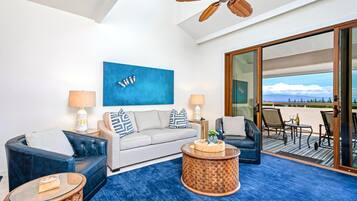 Un salon moderne comprenant un canapé, deux fauteuils, une table basse et un ventilateur de plafond. Une grande fenêtre offre une vue sur la plage.