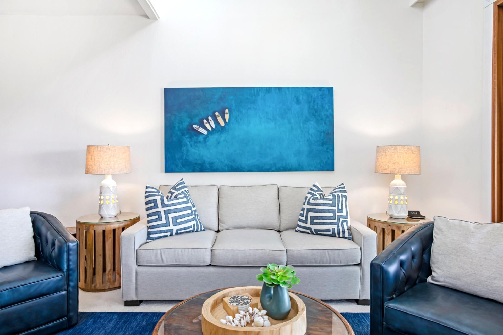 Ein modernes Wohnzimmer mit einem grauen Sofa, zwei Beistelltischen mit Lampen, einem Couchtisch mit Dekoration und einem blauen abstrakten Gemälde an der Wand.
