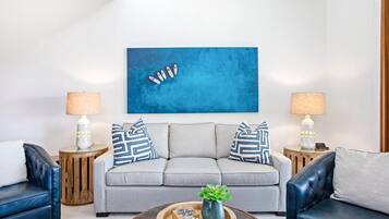 Ein modernes Wohnzimmer mit einem grauen Sofa, zwei Beistelltischen mit Lampen, einem Couchtisch mit Dekoration und einem blauen abstrakten Gemälde an der Wand.