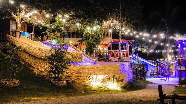 Een terras in de avond met een stenen muur, lichtjes en een bord met de tekst 'Into the Midst'.