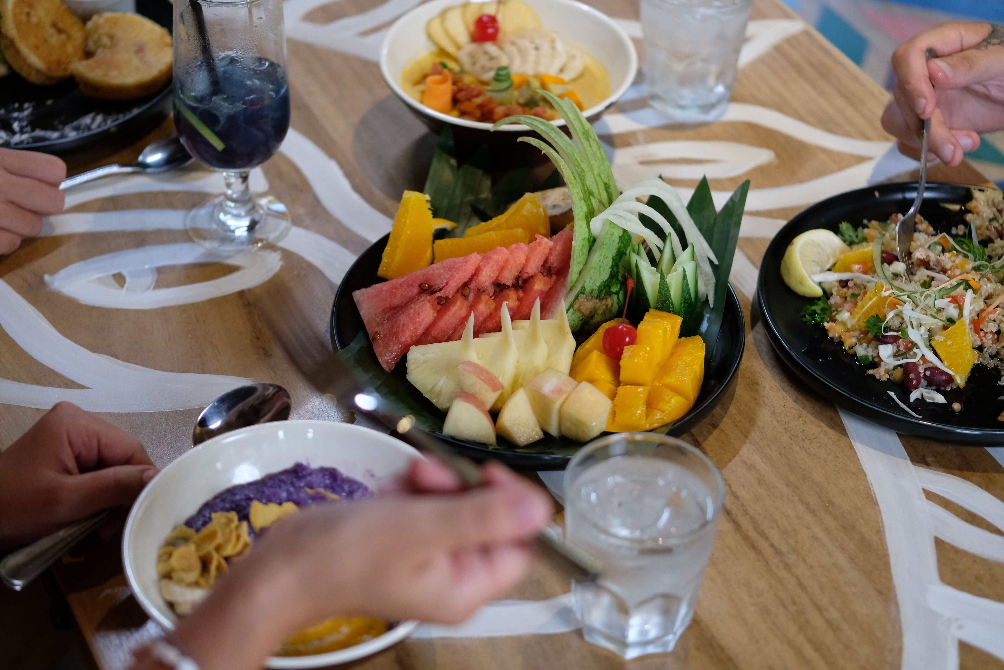 A table with various dishes including watermelon, mango, and a salad, with people dining.