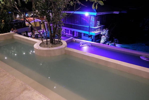 A swimming pool with a tiled deck, surrounded by trees and a building with a balcony.