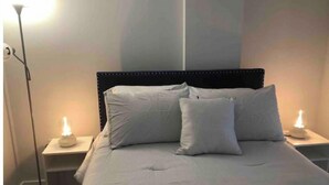 Un llit ben fet amb un capçal, dues làmpades laterals i una làmpada de peu en una habitació amb parets sense decoració.