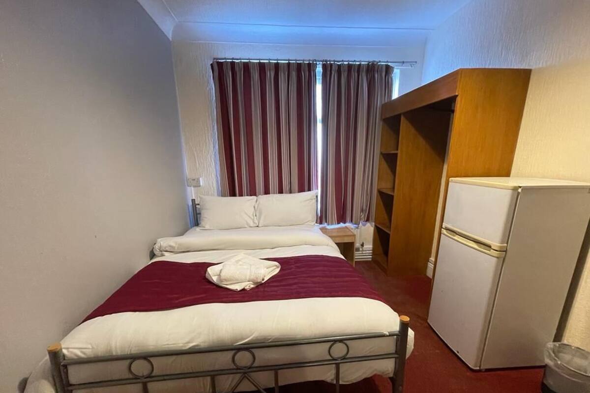 A small, single-bed room with a metal frame, a white refrigerator, and a wooden wardrobe.