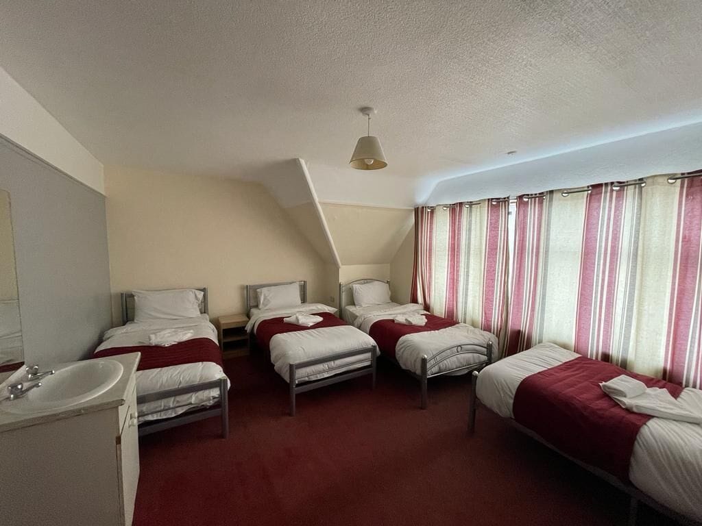A hotel room with four single beds, a small bathroom with a sink, and a window with striped curtains.