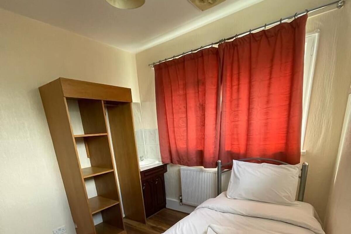 A bedroom with a bed, a wooden wardrobe, a radiator, and a window with red curtains.