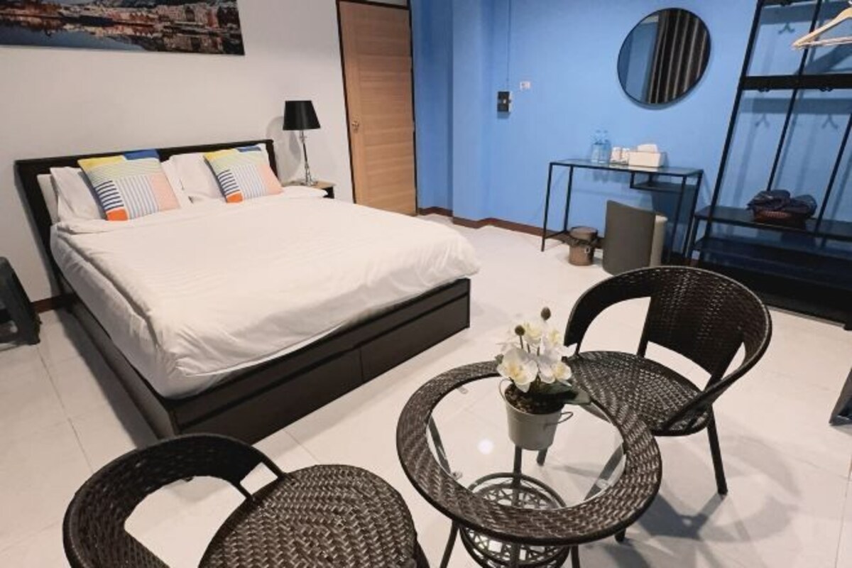 A hotel room with a bed, two wicker chairs, a small table with a potted plant, a mirror, a side table with a lamp, and a blue wall.