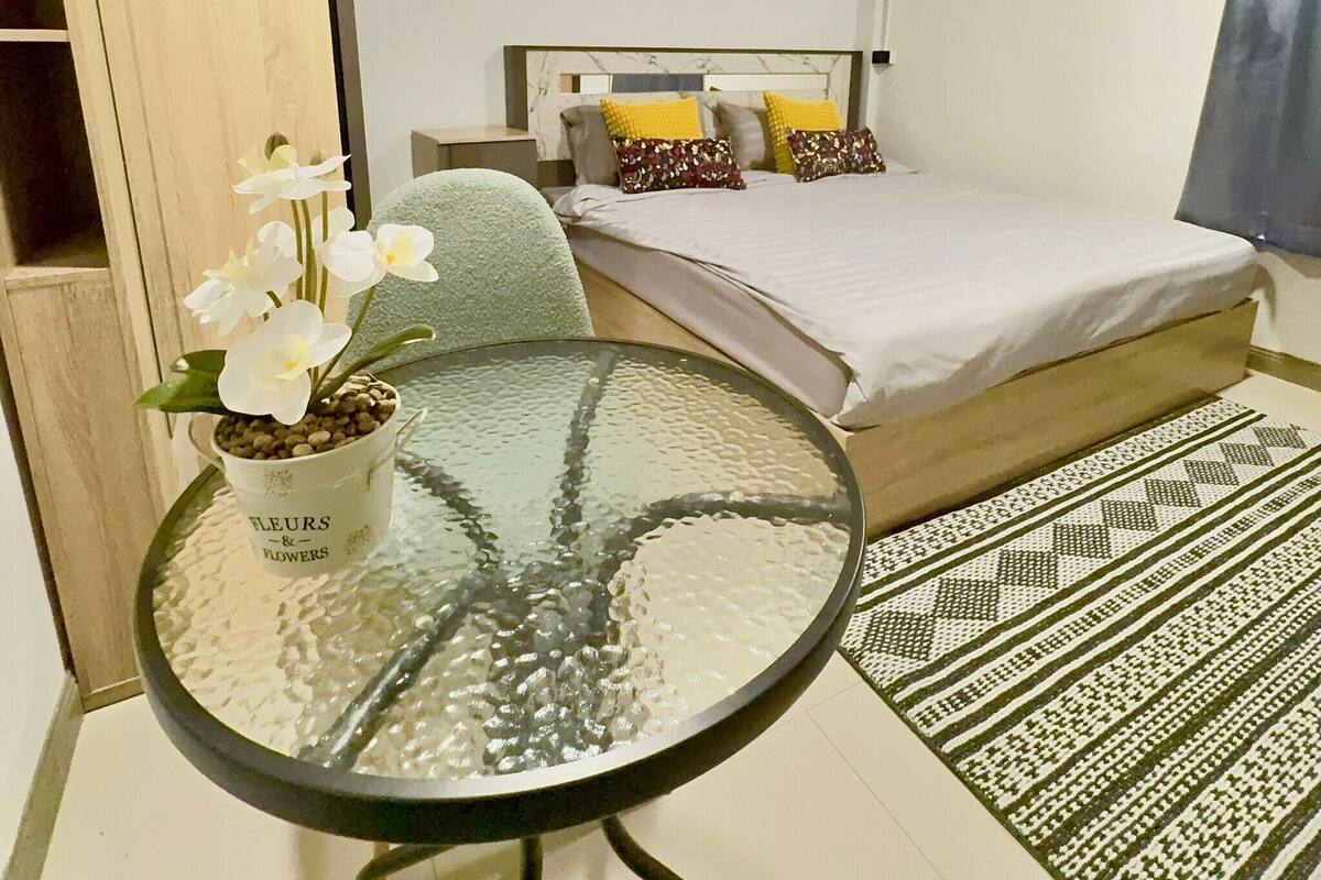 A bedroom with a bed, a bedside table with a potted plant, and a patterned rug.
