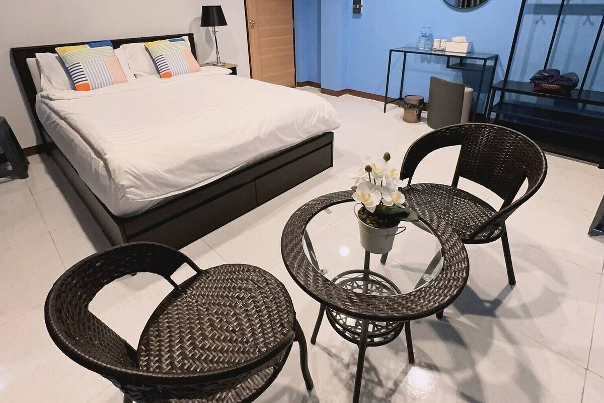 A modern bedroom with a large bed, two wicker chairs, a small table with flowers, and a view of the city through a window.