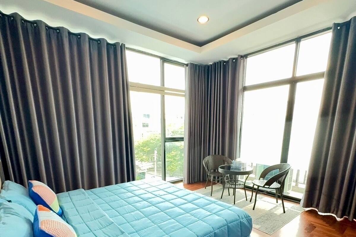 A hotel room with a large bed, a small table with chairs, and large windows with curtains.