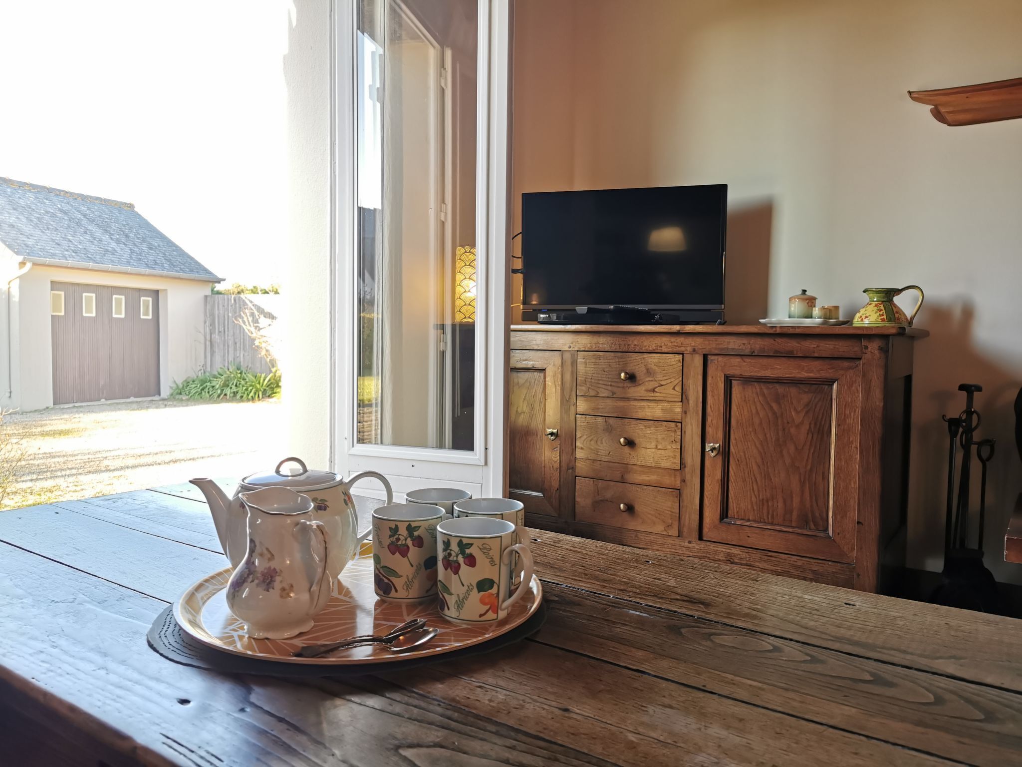 A wooden table with a tray of teapots and cups, a television on a wooden cabinet, and a window with a view of a house and fence.