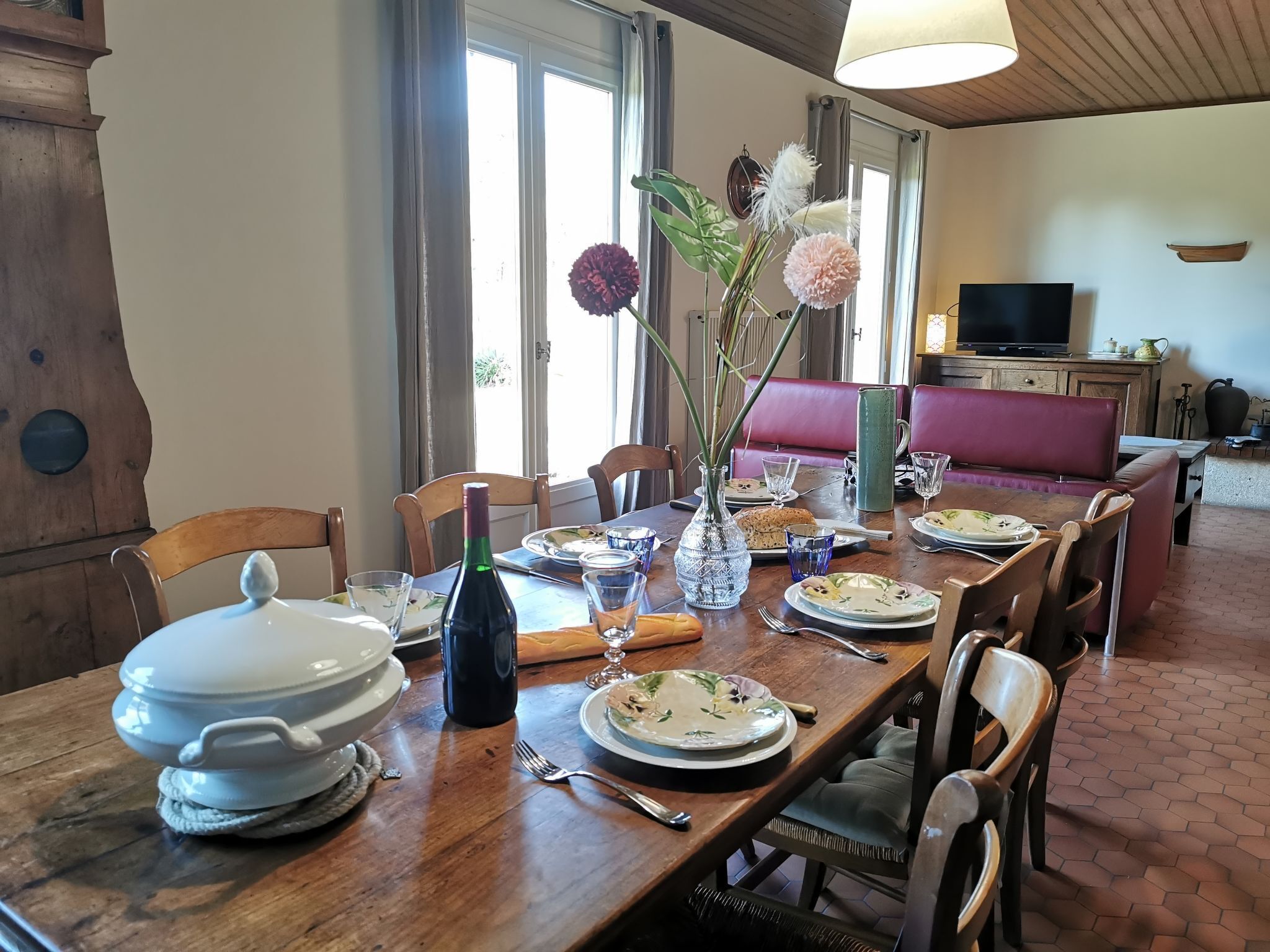 Un comedor con una mesa de madera dispuesta para una comida, un jarrón con flores y un televisor al fondo.