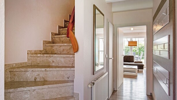 A staircase with a wooden handrail leading to a room with a large mirror, a radiator, and a view of a living area with a sofa and a hanging light fixture. - MeerZeit I by Interhome (Burhave)