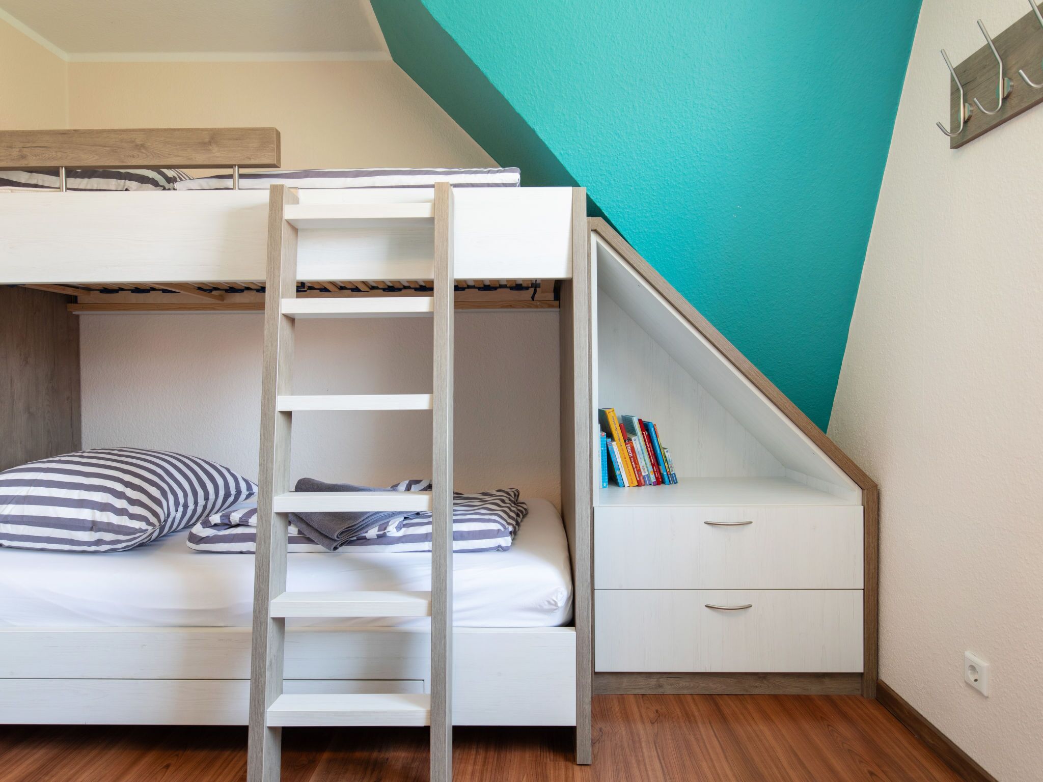 A bunk bed with a ladder, a small desk with drawers, and a bookshelf under a slanted ceiling.