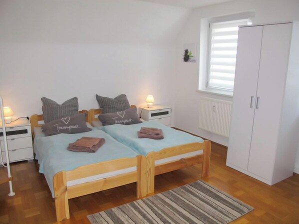 Un dormitorio con dos camas de madera, mesitas de noche blancas, un armario blanco y una ventana con persianas.