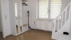 A room with a wooden floor, white walls, a white radiator, a window with blinds, and a staircase.