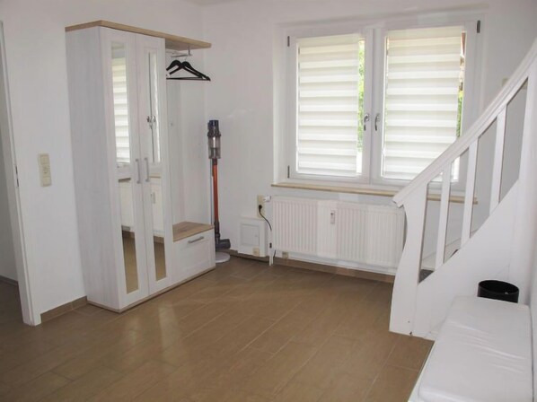 A room with a wooden floor, white walls, a white radiator, a window with blinds, and a staircase.