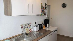 A compact kitchen with white cabinets, a stainless steel sink, a coffee maker, and a clock on the wall.