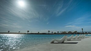 A beach with sunlit water, white lounge chairs, palm trees, and a building with a unique roof structure. - Joya Vacation Club (Vítor)