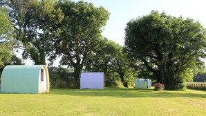 Three small, rectangular, covered structures in a grassy field with trees in the background. - The Pod Garden- Cozy Glamping Pod, Rural Location (Newquay)