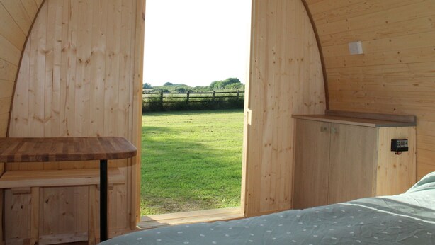 A wooden interior with a bed, a small table, and a view of a grassy field with a fence. - The Pod Garden- Cozy Glamping Pod, Rural Location (Newquay)