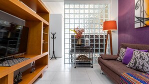 A modern living room with a TV on a wooden stand, a sofa with patterned cushions, a floor lamp, and a decorative shelf with a vase. - Modern Studio Next to Arpoador Jc1012 Z2 (Rio de Janeiro)