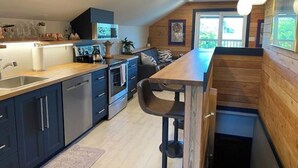 Une cuisine moderne avec des meubles de couleur sombre, un plan de travail en bois et un four encastré. On y trouve un coin repas avec une table et des chaises en bois, ainsi qu’une grande fenêtre qui laisse entrer la lumière naturelle.