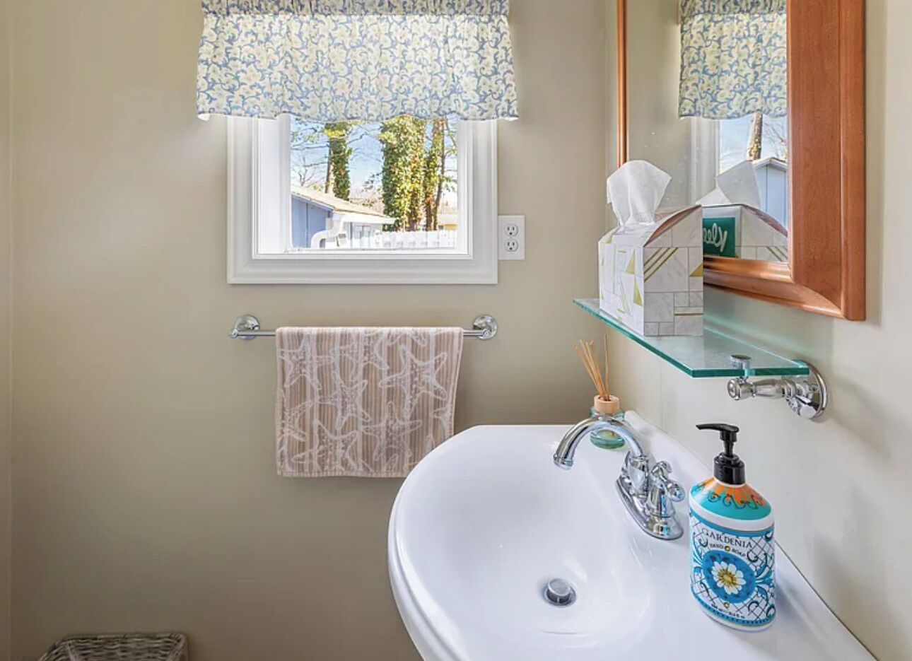 Un baño con un lavamanos blanco, un estante de vidrio, un perchero con una toalla estampada y una ventana con vistas a la vegetación.