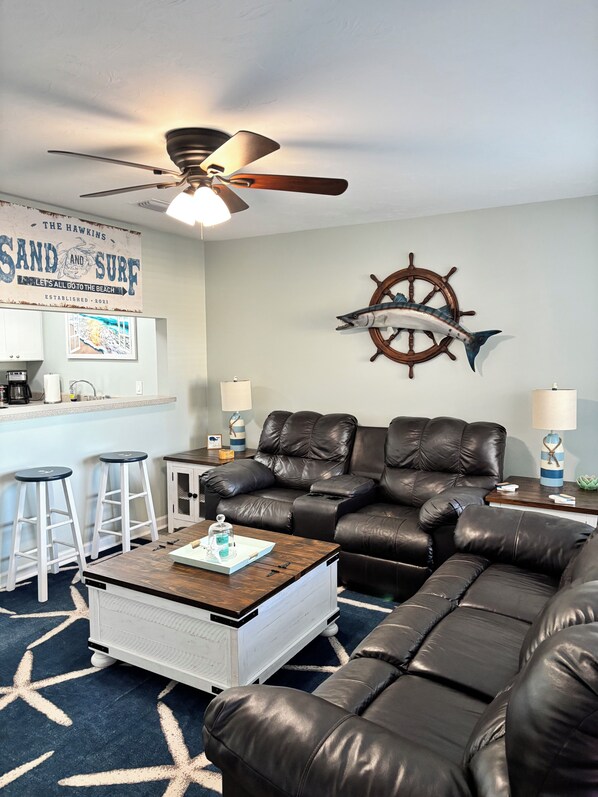 A living room with a black leather sofa, a wooden coffee table, a ceiling fan, a ship's wheel decoration, and a kitchen area with bar stools.