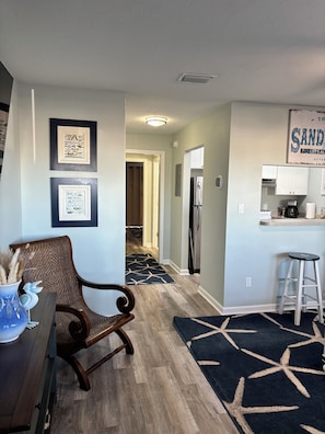 A living room with a dining table, a chair, a kitchen area with a refrigerator, and a hallway leading to another room.