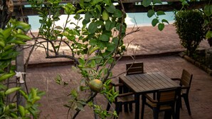 Um pátio externo com mesa e cadeiras de madeira, piso de tijolos e vista para uma piscina.