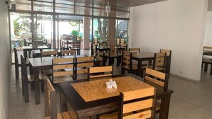 Uma sala de jantar ampla e interna, com várias mesas e cadeiras de madeira, janelas grandes que permitem entrada de luz natural e um design simples e clean.